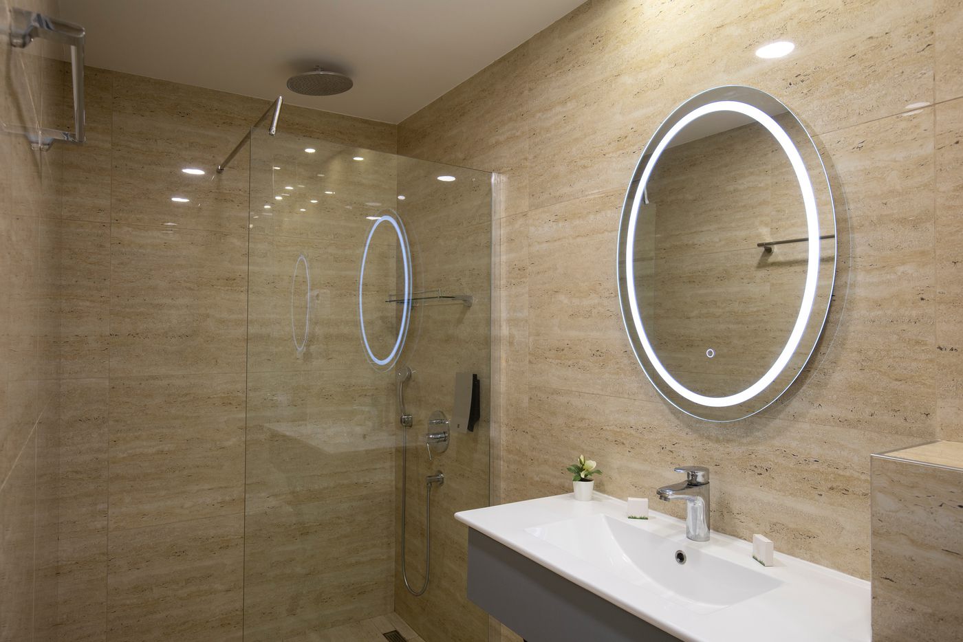 Hotel bathroom with warm beige stone-look wall surfaces, a glass shower, a backlit round mirror and a white basin